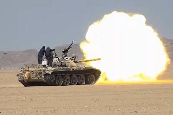 Un char tir sur une force armée opposée au mileu d'une zone aride. Le tir provoque une énorme boule de feu jaune au bout du canon du char.