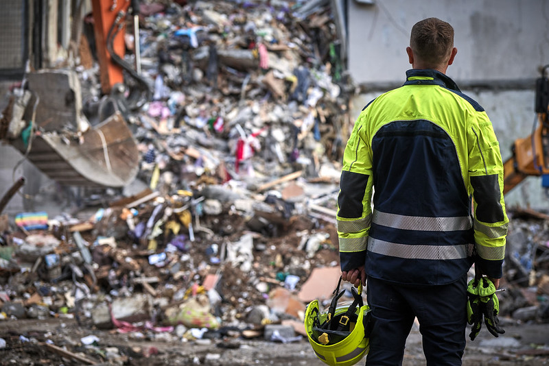 Une personne, de dos, avec un uniforme jaune fluo et bleu foncé, regarde des débris de ce qui semble être un bâtiment.