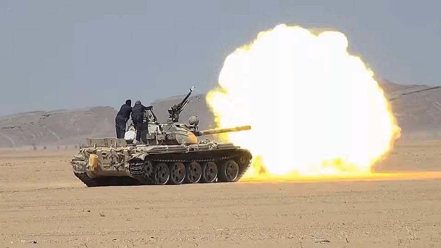Un char tir sur une force armée opposée au mileu d'une zone aride. Le tir provoque une énorme boule de feu jaune au bout du canon du char.