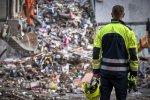 Une personne, de dos, avec un uniforme jaune fluo et bleu foncé, regarde des débris de ce qui semble être un bâtiment.